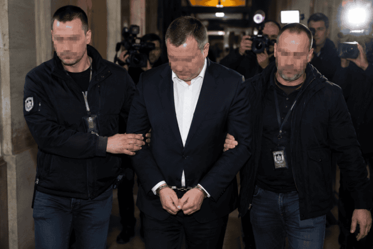 Bilincsben vezetnek egy öltönyös politikust egy hivatali épület folyosóján, újságírók és kamerák előtt.